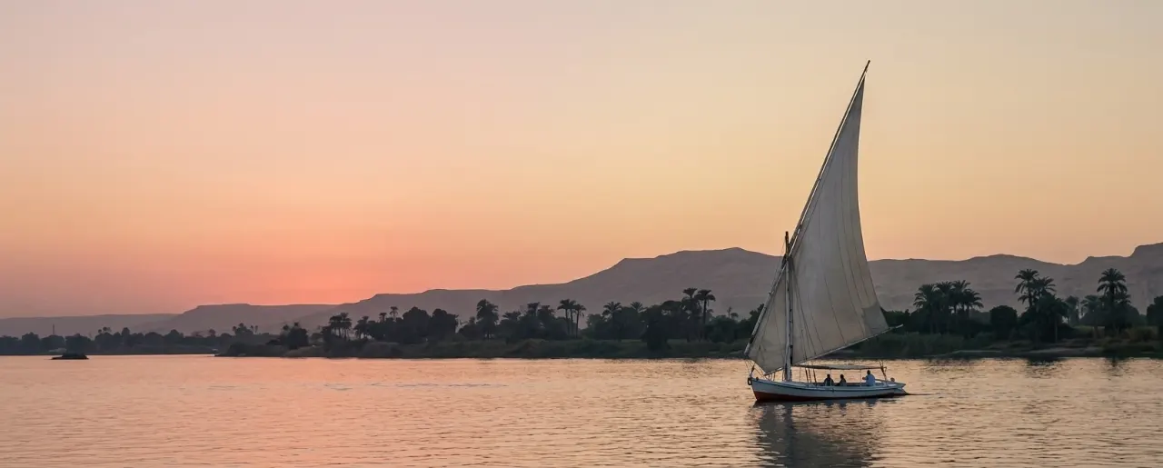 A felucca under full sail on the Nile at sunset, palms and the desert hills of the west bank behind