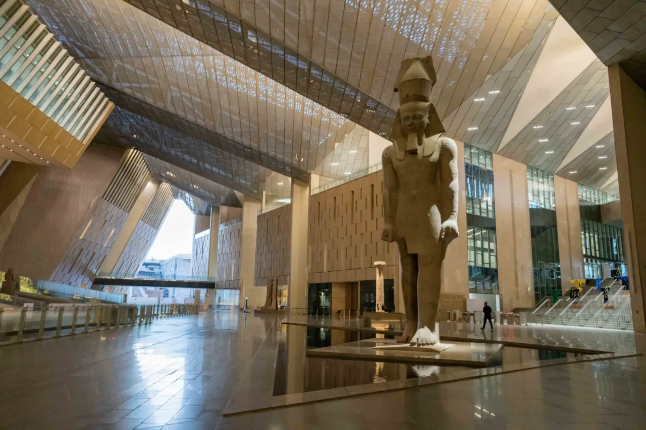 The colossal statue of Ramesses II above a still pool in the atrium of the Grand Egyptian Museum, a single visitor crossing the hall