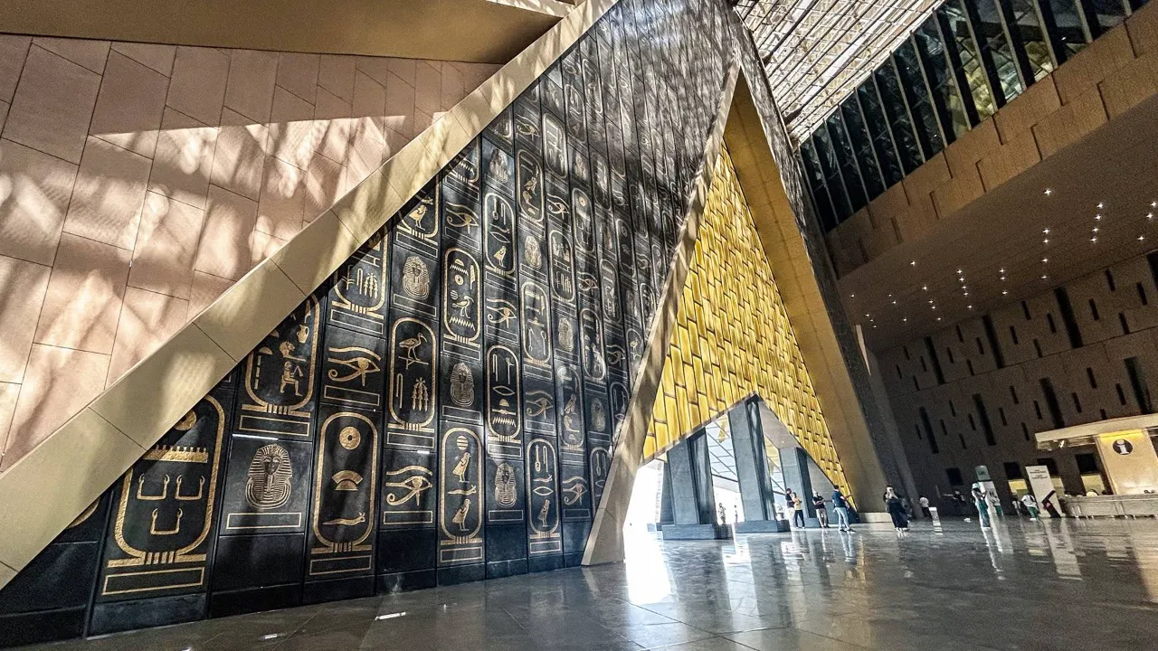A wall of gilded royal cartouches on black stone inside the Grand Egyptian Museum, light falling across it beside a golden pyramidal entrance