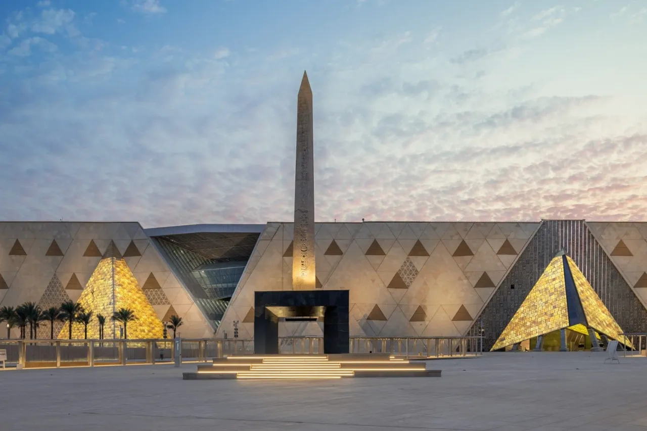 The obelisk of Ramesses II in the forecourt of the Grand Egyptian Museum at dusk, the lit faÃ§ade behind