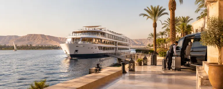 A driver loading luggage into a car on a riverside quay as a Nile ship comes alongside