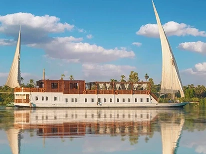 A dahabiya under twin sails on the Nile in daylight, reflected in still water beneath palm-lined banks