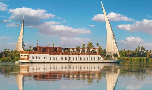 A dahabiya under sail on the Nile, twin sails raised above a wooden upper deck