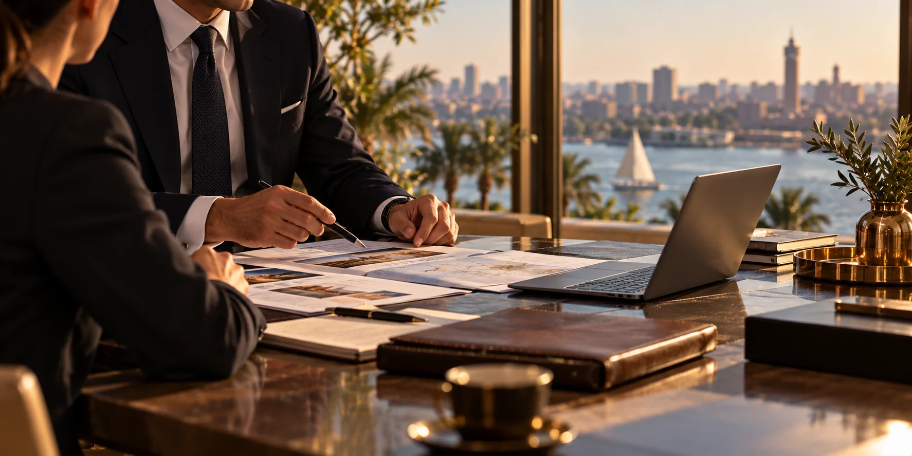 Two people in dark suits go through printed itineraries at a desk, a laptop beside them and a felucca on the Nile through the window